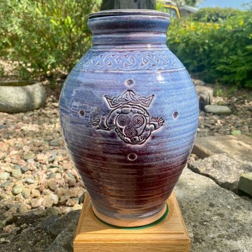 Purple heather cremation Urn on plinth with greenery behind it